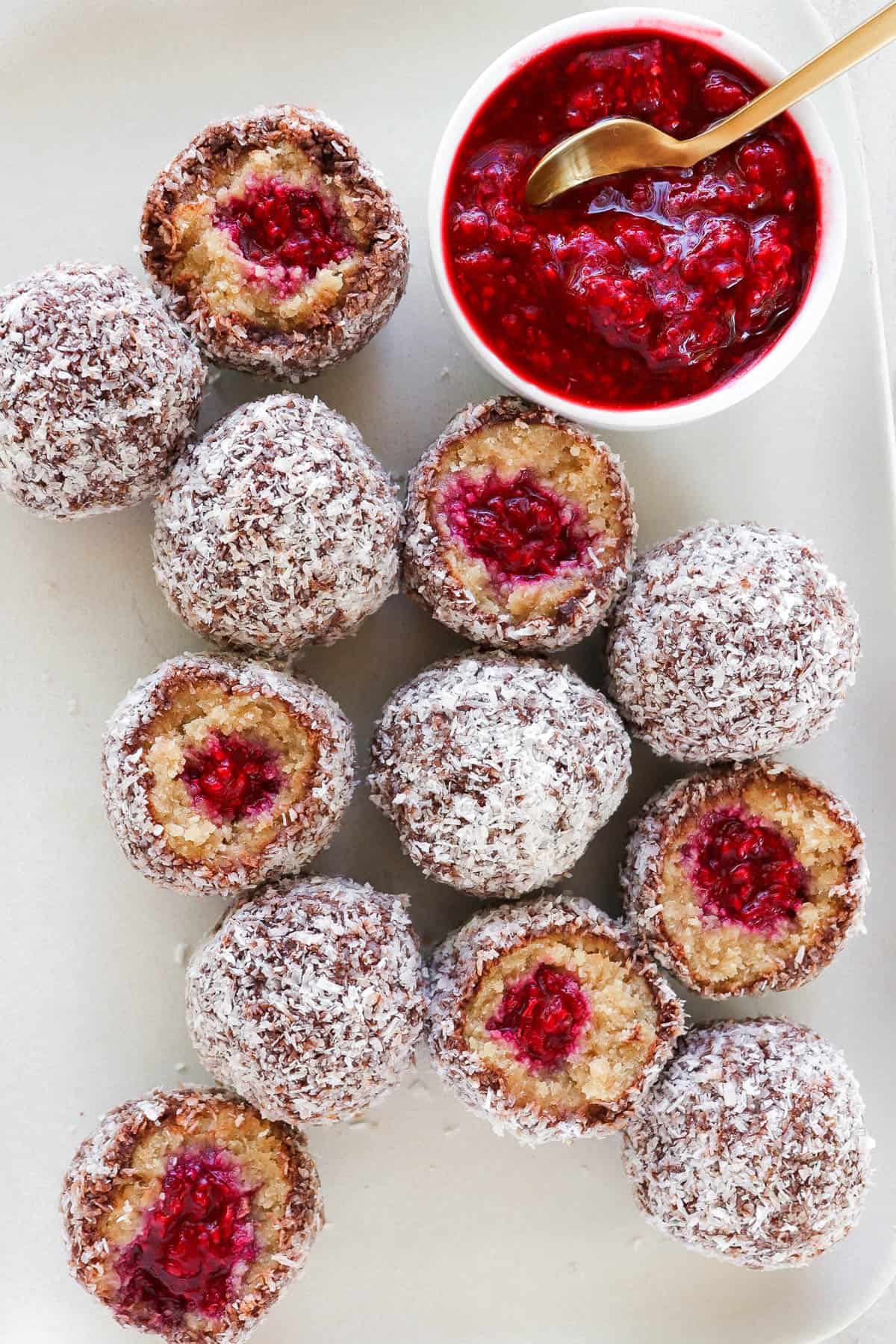 Plate of Lamington balls cut in half. Mashed raspberries on side with gold spoon for decoration.
