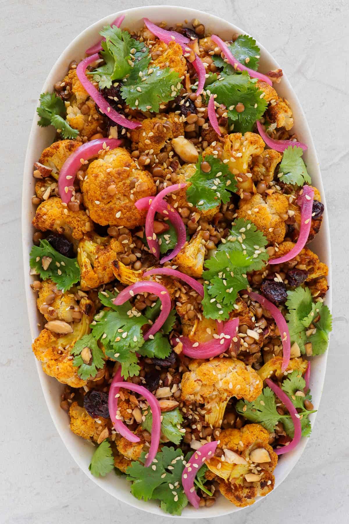 Roasted cauliflower on a plate with coriander leaves, pickled onions, lentils, dried cranberries and toasted sesame seeds.
