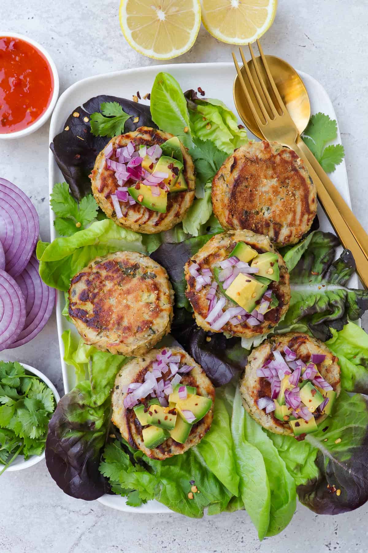 Tuna cakes on lettuce leaves with chopped avocado, red onion and chili flakes on top of some. Sliced red onion, coriander leaves, sweet chilli sauce and lemons on the side for decoration.