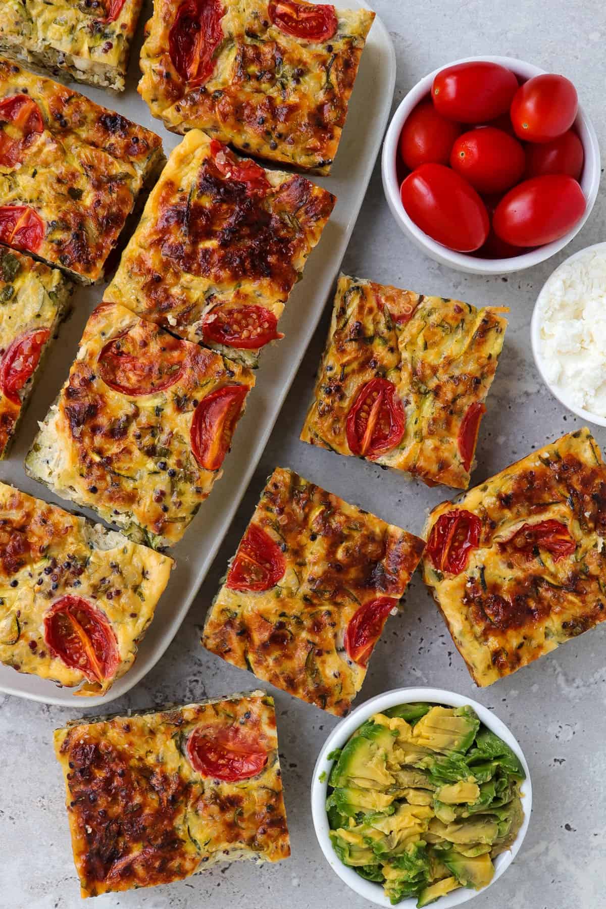 Some cut up zucchini slice squares on a plate and some on the table. Small dishes of cottage cheese, mashed avocado and cherry tomatoes for decoration.