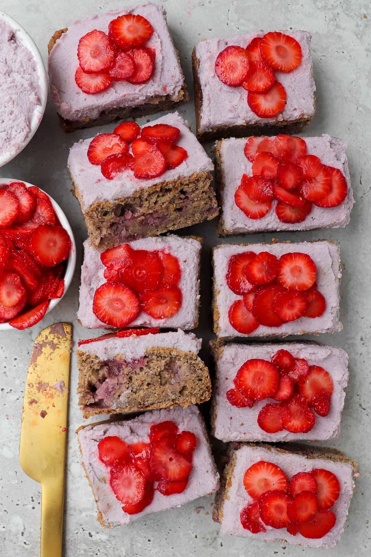 Frosted cake topped with sliced strawberries and cut up into small rectangle slices. two slices tilted on the side or an inside view.