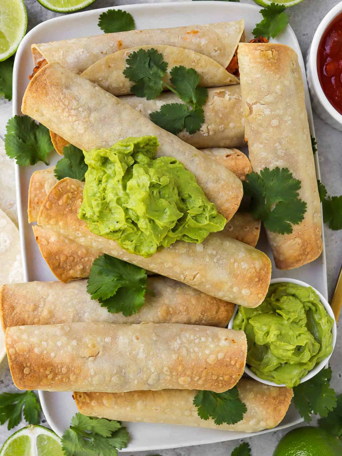 Taquitos on platter topped with coriander leaves and guacamole. Fresh cut up limes, tortillas, salsa and gold spoon on the sides.
