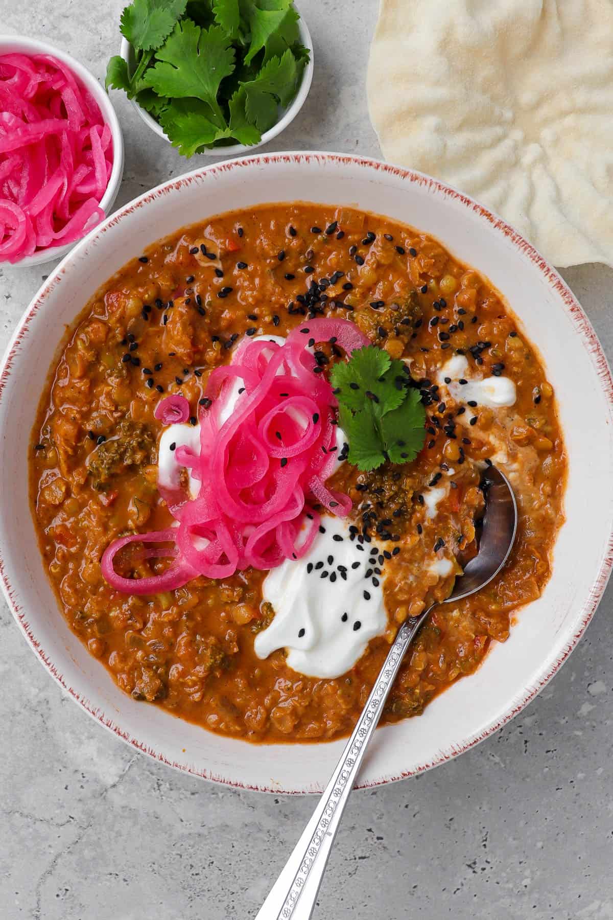 Dahl served in a serving bowl with spoon. Topped with pickled onions, yoghurt, coriander leaves and black sesame seeds for garnish.