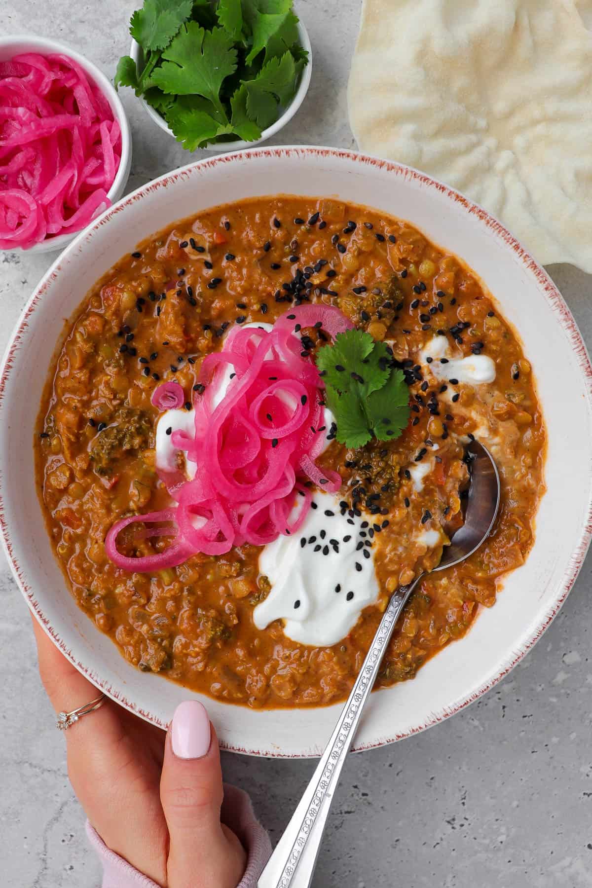 Holding bowl of dahl with spoon. Topped with pickled onions, yoghurt, coriander leaves and black sesame seeds for garnish.