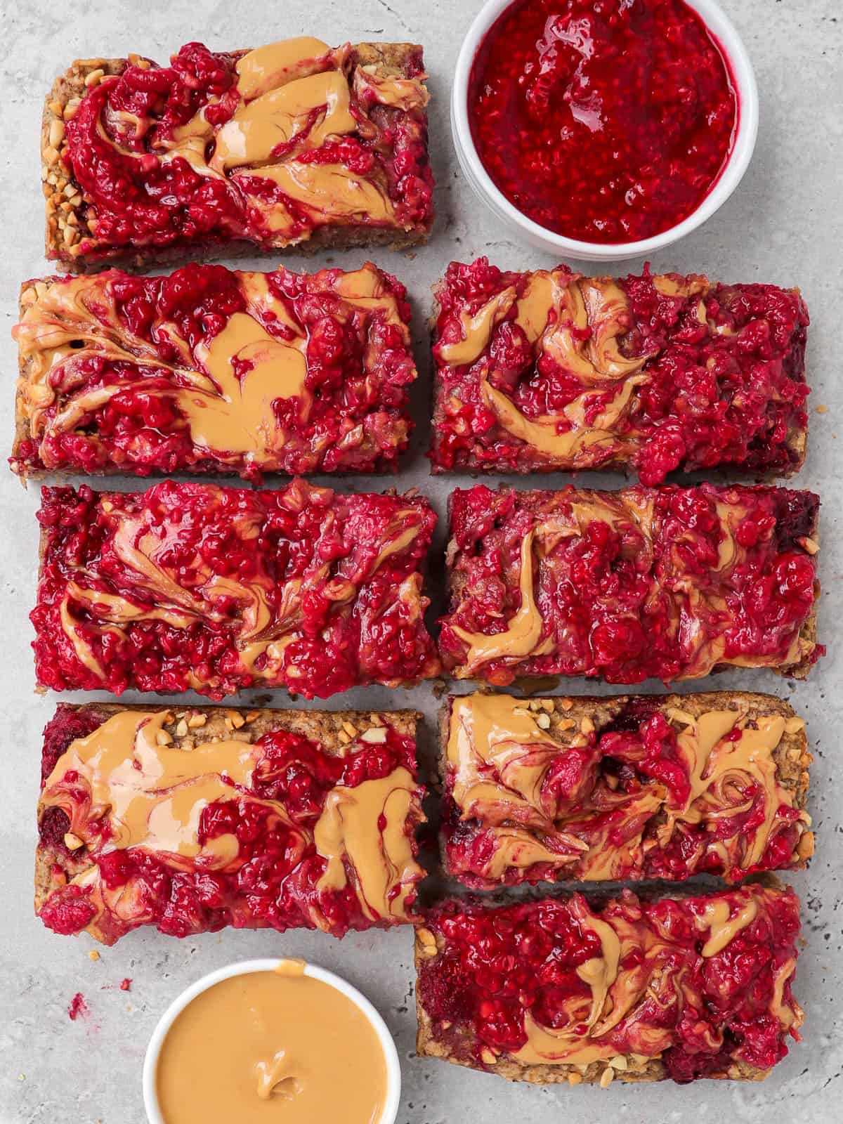 Peanut butter and jelly oatmeal cut up into bars. Small dish of peanut butter and mashed raspberries at top and bottom.