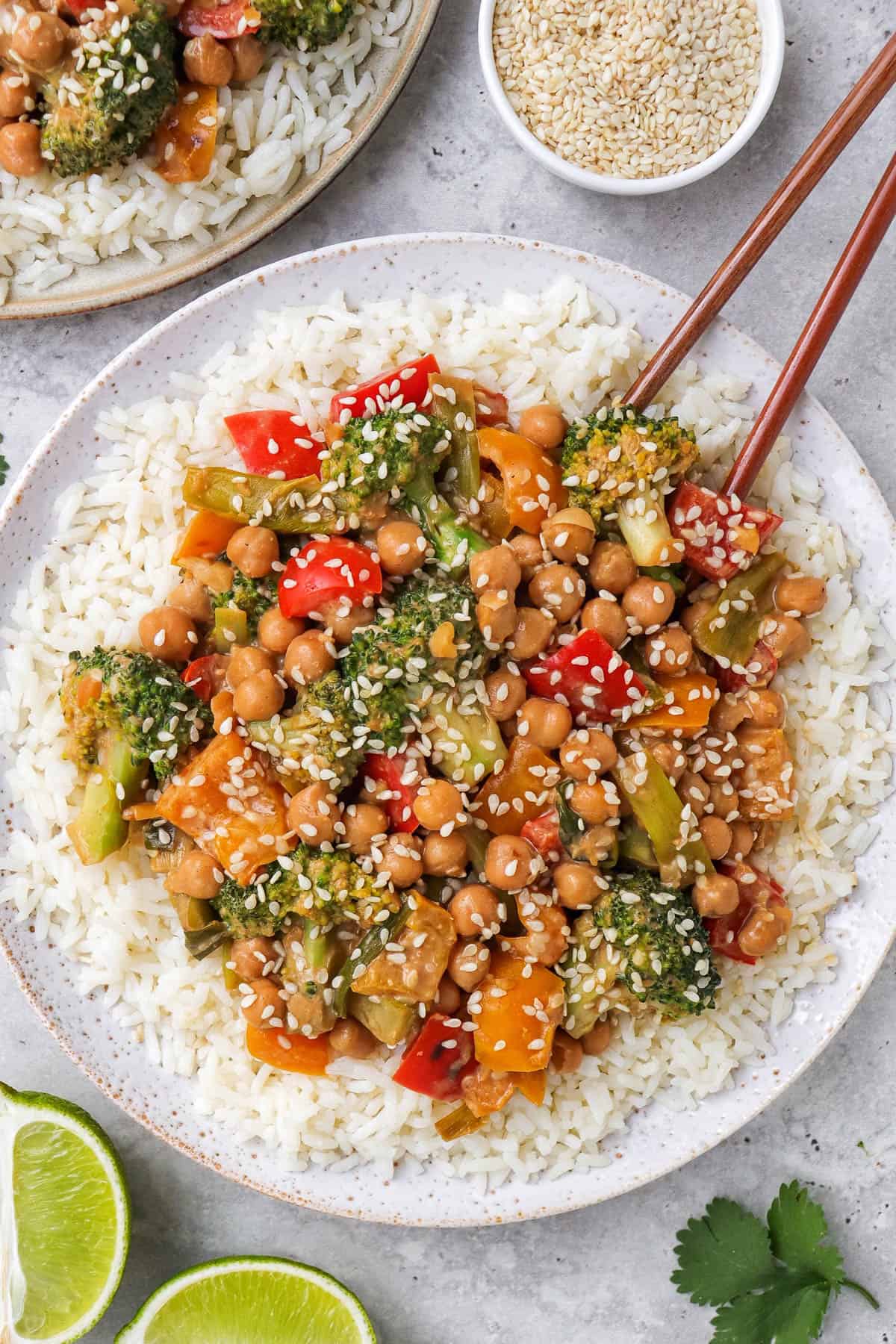 Chickpea vegetable stir fry on a plate with rice and chop sticks. Sesame seeds and cut up limes on the side.