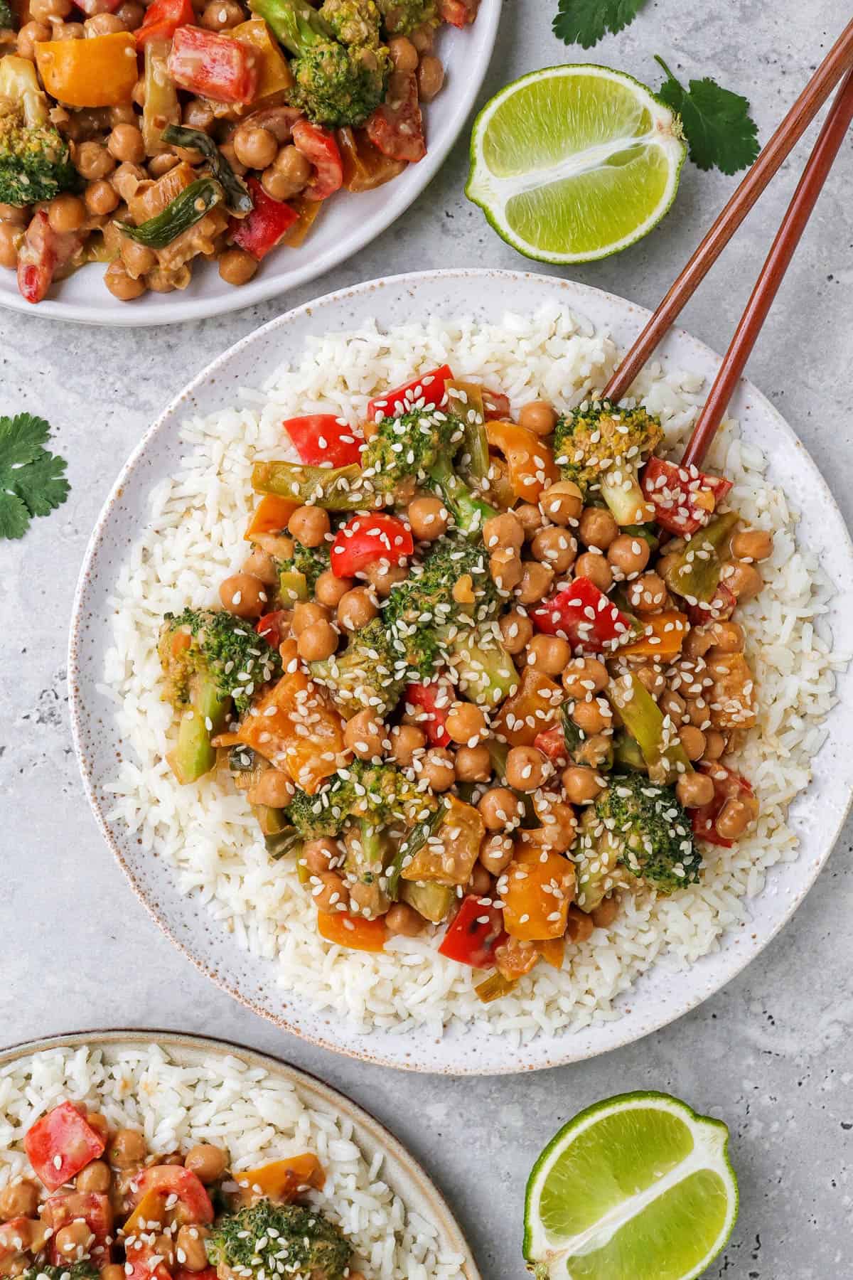 Chickpea vegetable stir fry on 3 plates with rice and chop sticks. Sesame seeds and cut up limes on the side.