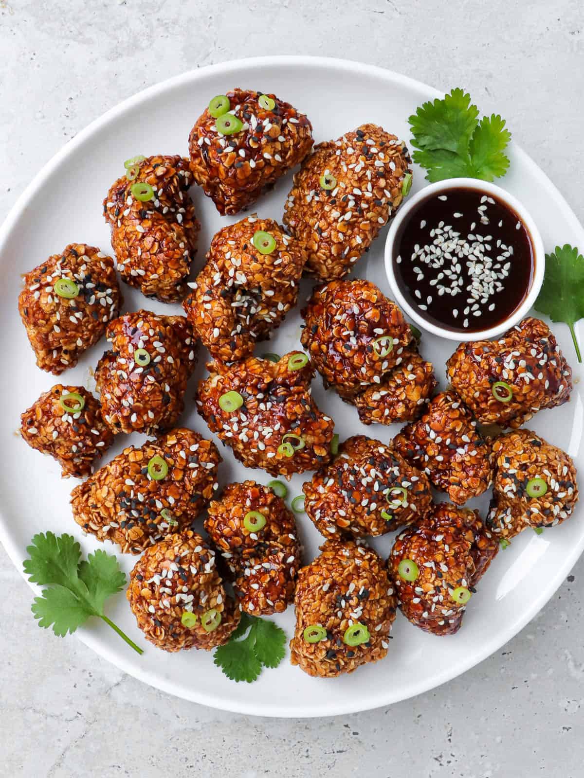 Teriyaki cauliflower wings topped with spring onions on a plate with coriander leaves and sauce on side.