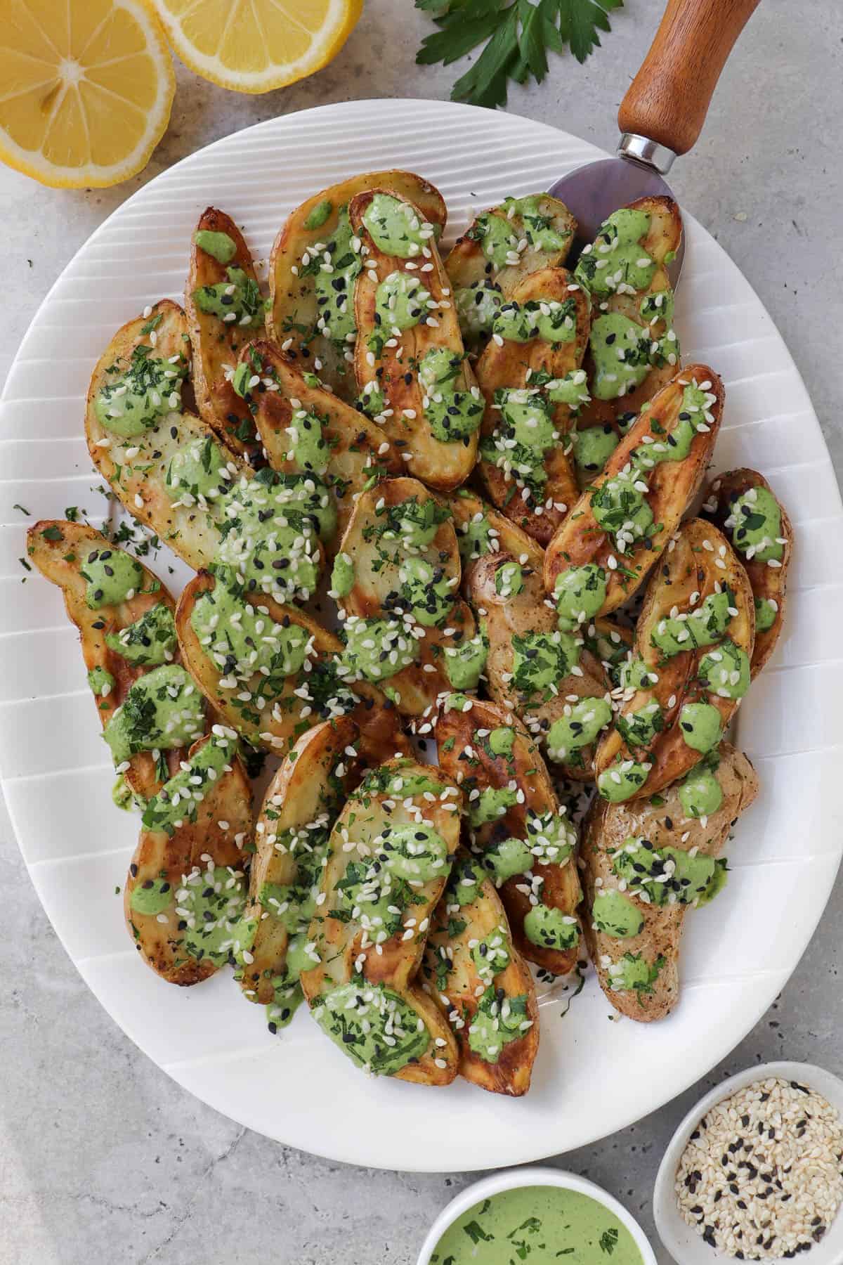 Air fryer fingerling potatoes on plate with green tahini sauce. Lemons, sauce, sesame seeds and parsley on the side.
