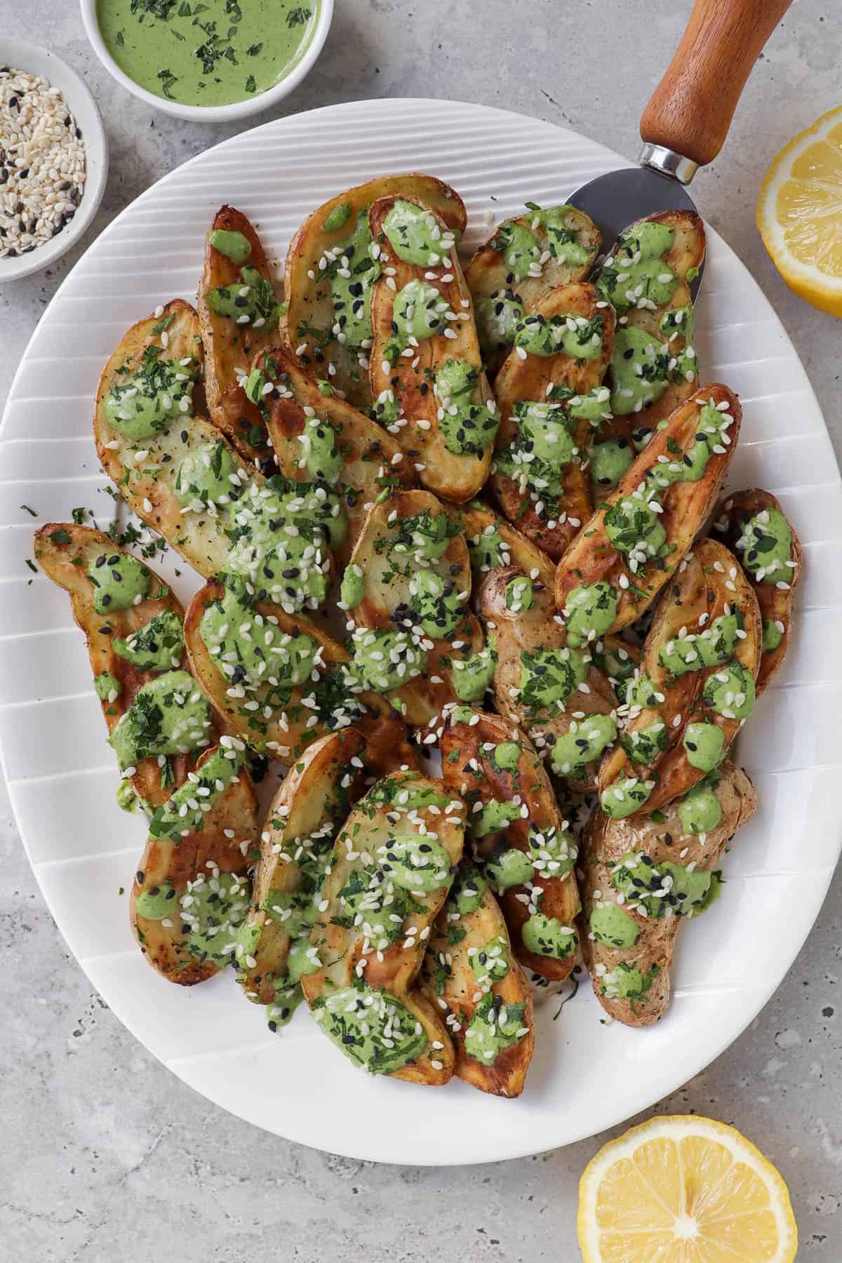 Air fryer fingerling potatoes on plate with green tahini sauce and serving spoon.. Lemons, extra tahini sauce, sesame seeds on the side.