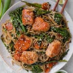 Broccolini, carrot soba noodle stir fry with chunks of crispy salmon on top. Chopsticks on plate.