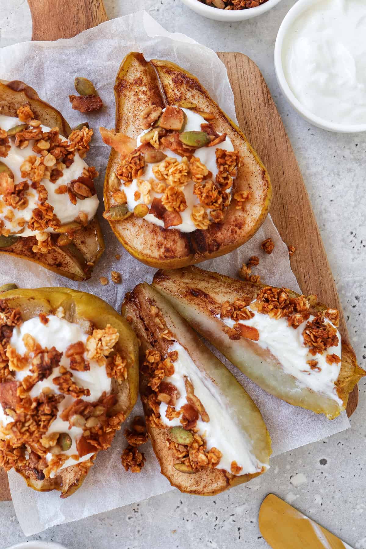 close up shot of baked pears on wooden board topped with plain yoghurt and granola. One pear cut in half.