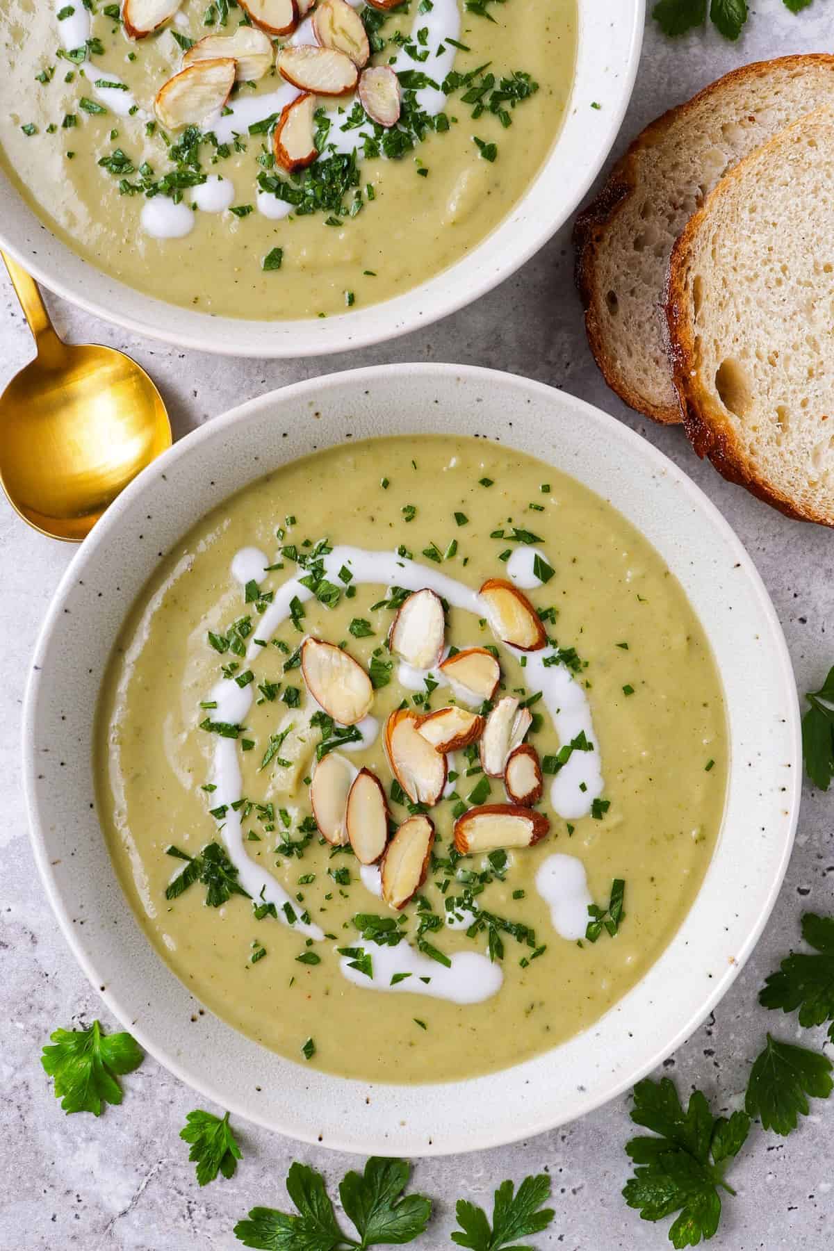 Broccoli soup in a bowl topped with coconut cream. almond flakes, finely chopped parsley. sourdough and golden spoon on the side.