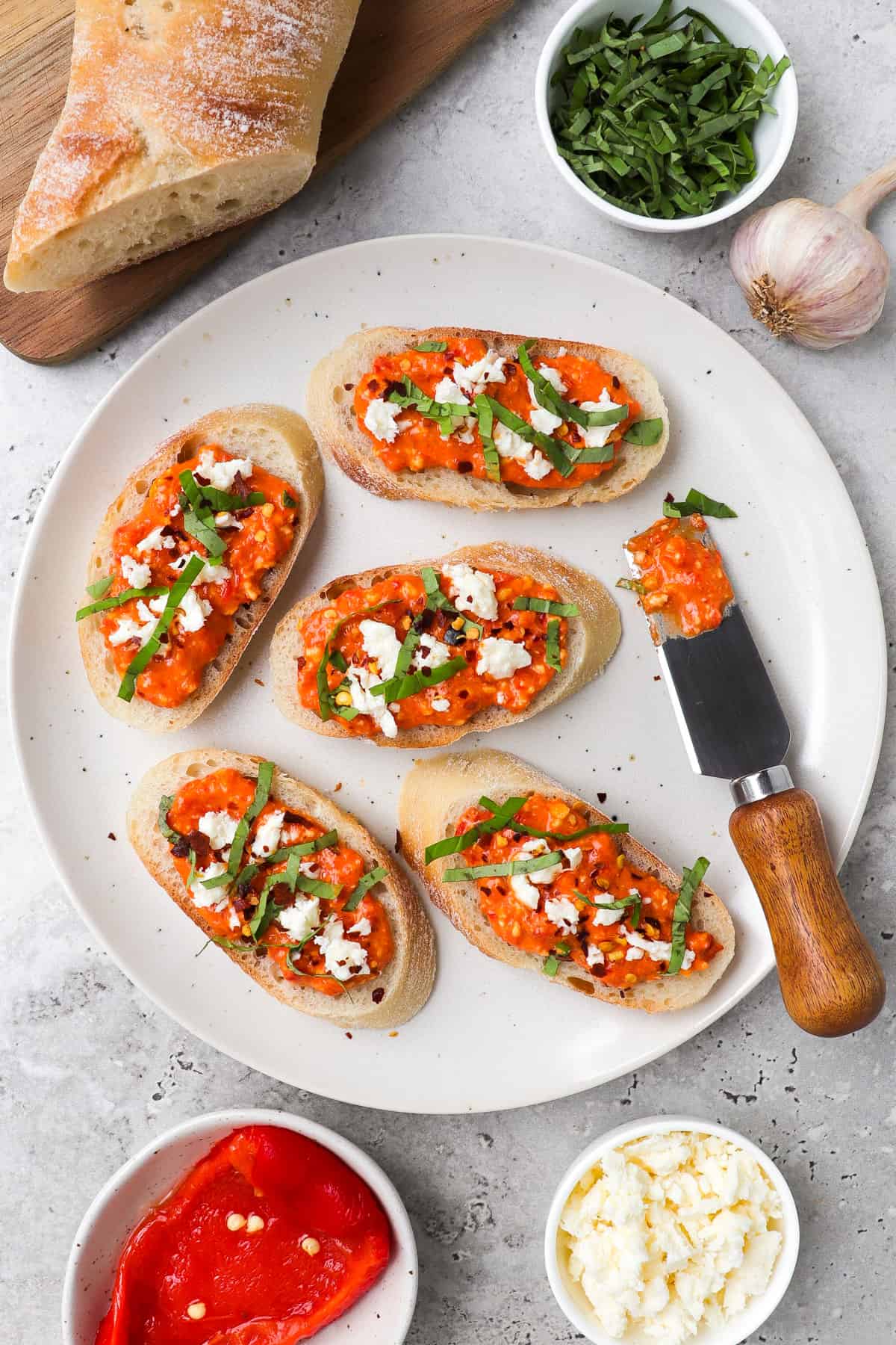 Crostini with paprika dip topped with feta, basil and chili flakes. Baguette, garlic, crumbled feta cheese, reasoned peppers on thee sides for garnish.