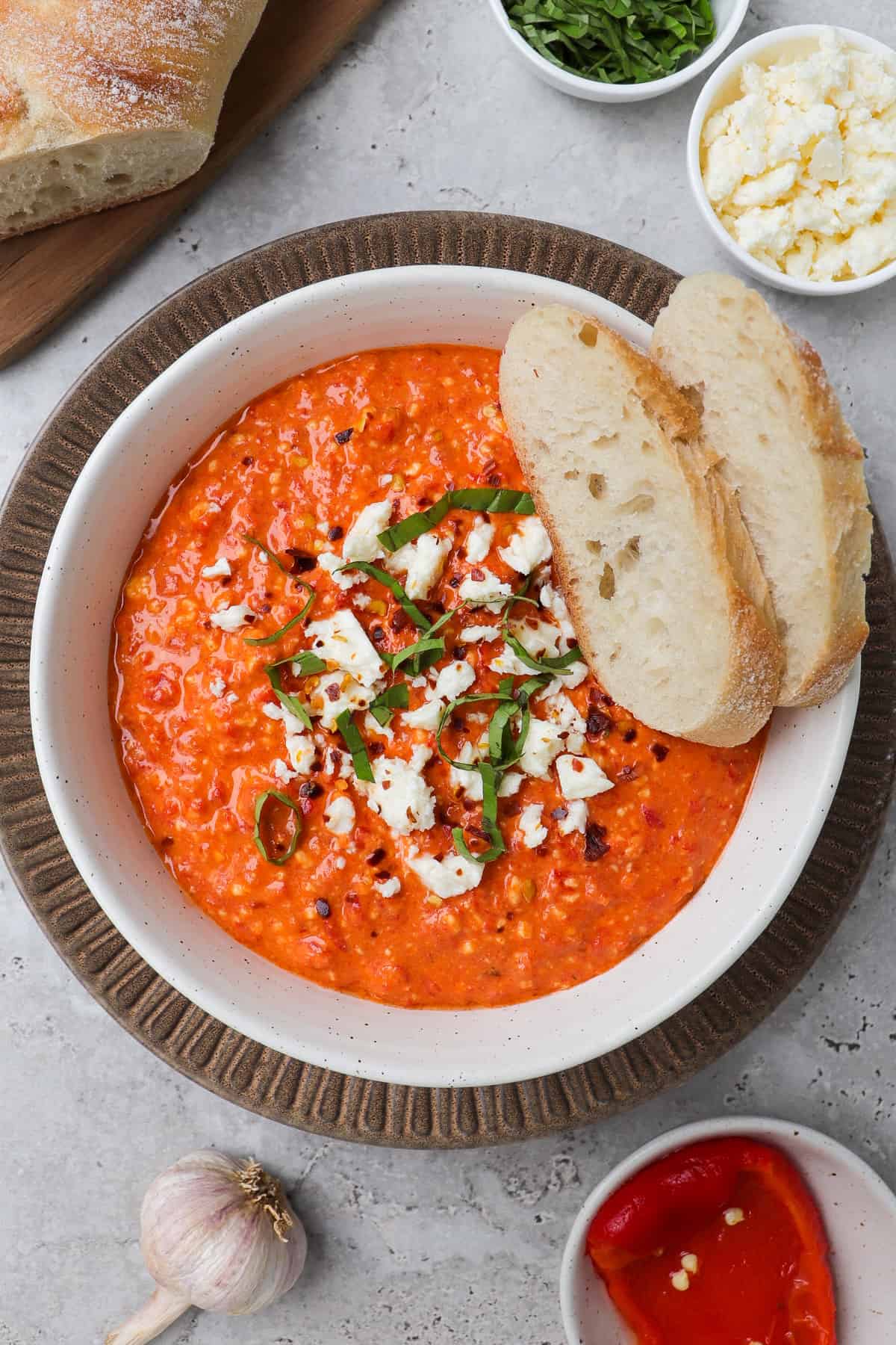 Bowl of paprika feta dip in a dish. Topped with crumbled feta, basil, chili flakes and sliced bread.
