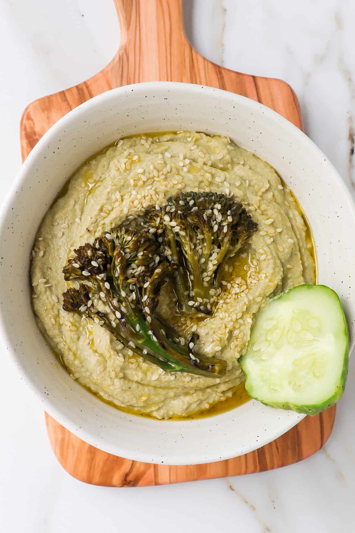 Hummus in a bowl topped with broccolini and cucumber chip.