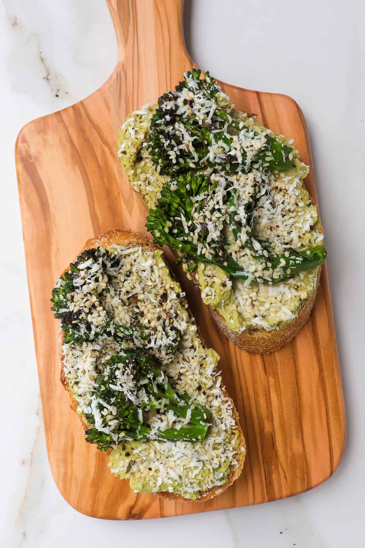 Cut up toast topped with broccoli hummus, broccolini and grated feta on wooden board.