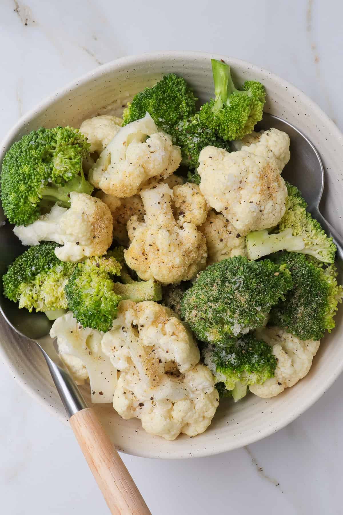 Raw broccoli and cauliflower florets in a bowl with seasonings and spoons.
