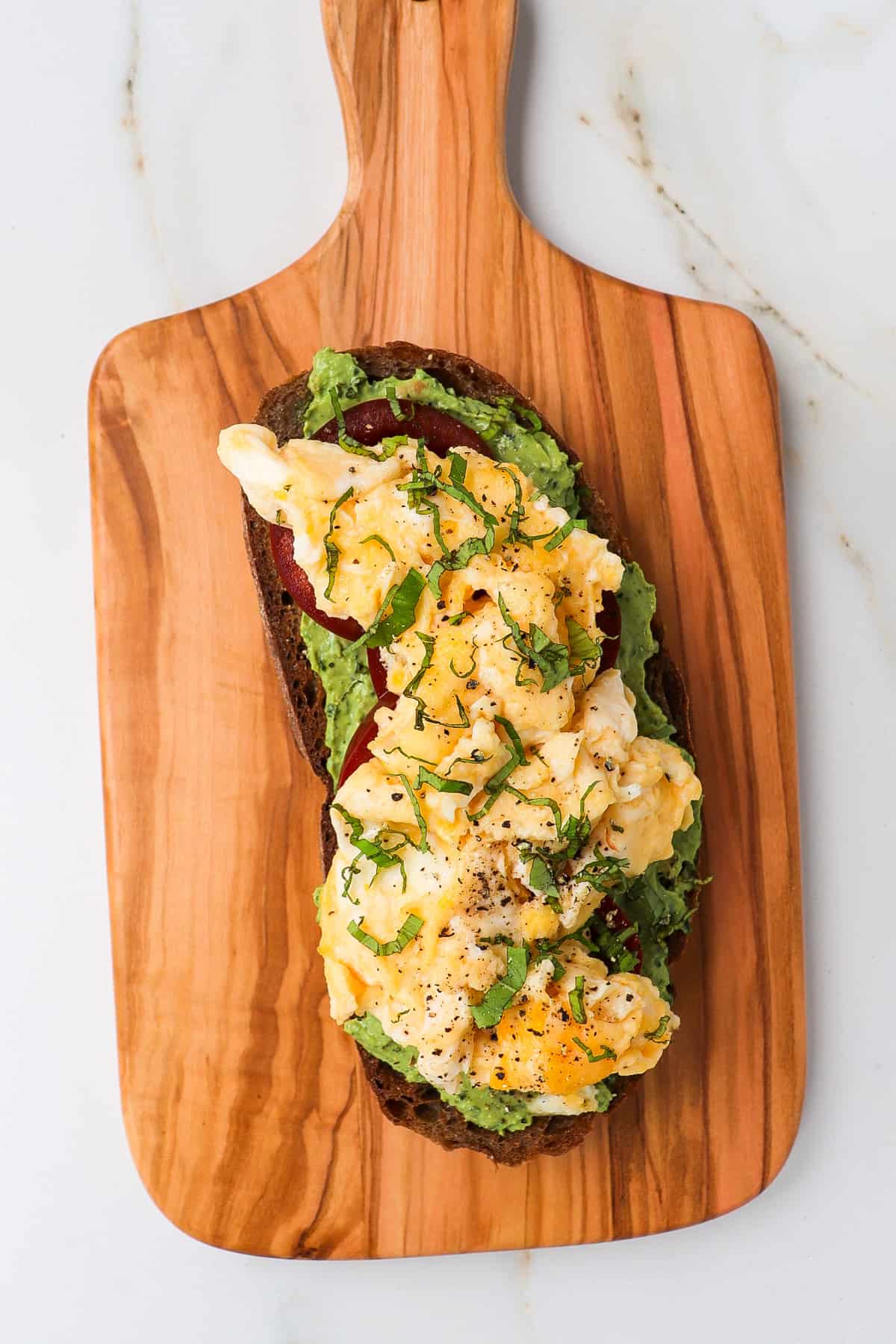 Slice of sourdough on a wooden board with pesto, scrambled eggs and chopped basil.