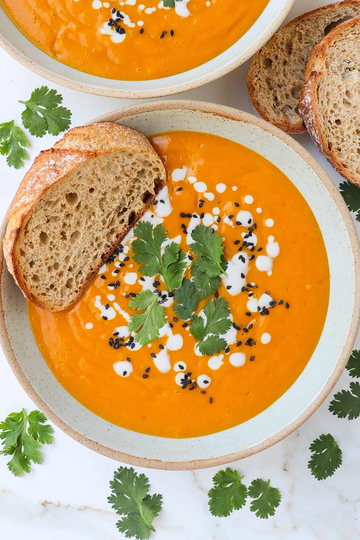 Sweet potato pumpkin soup in a bowl with coriander, coconut cream, sesame seeds and sliced bread on the side.
