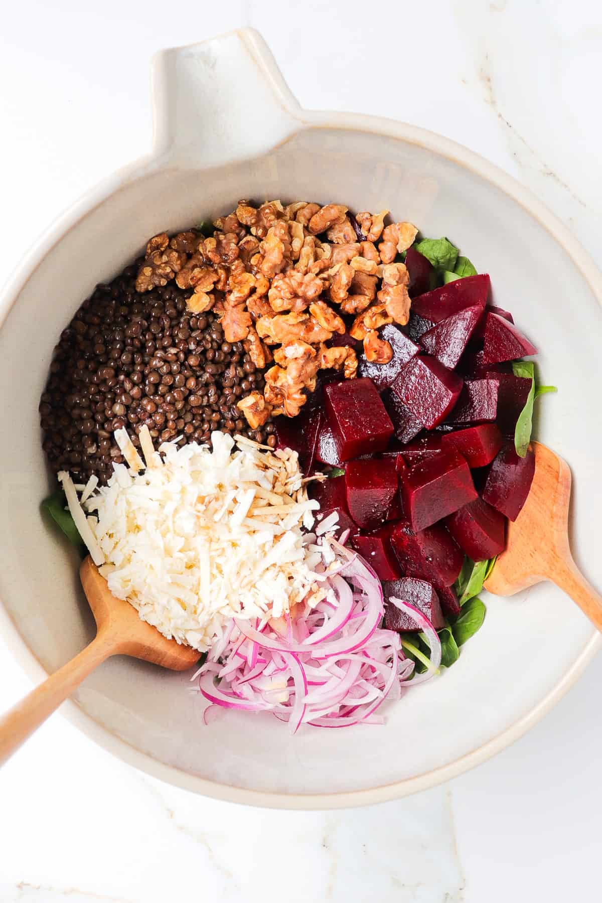 Spinach leaves, grated feta cheese, lentils, walnuts, beetroot and onions in a large mixing bowl with wooden spoons.