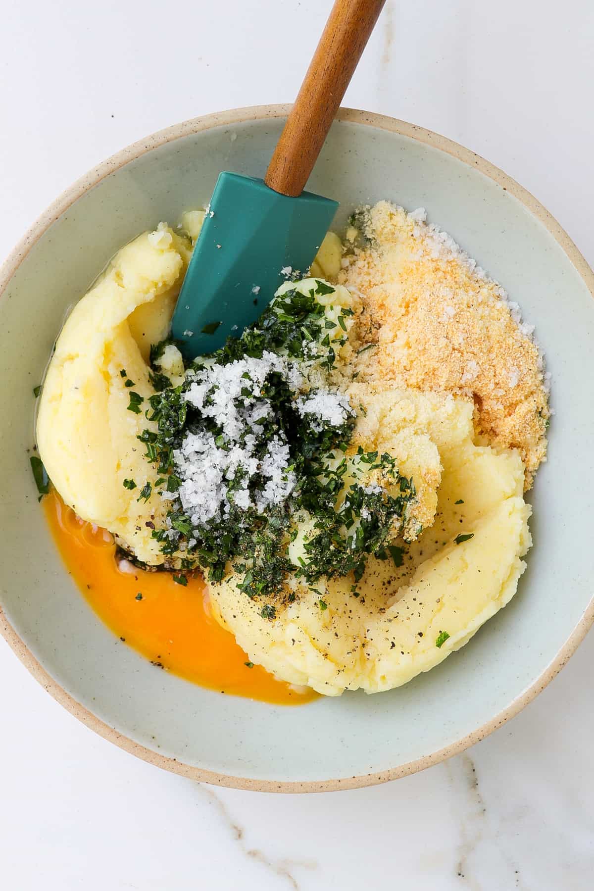 Mashed potatoes, egg, herbs and seasoning in a bowl with a spatula.