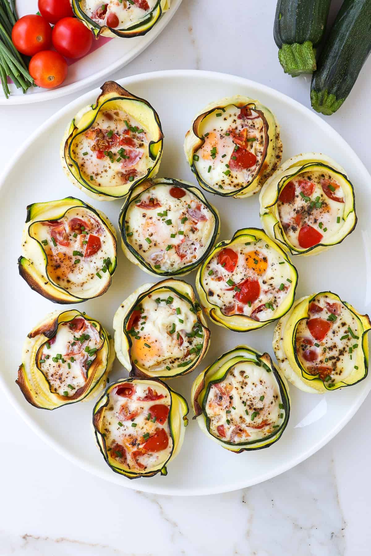 Zucchini wrapped egg cups on a plate with zucchini and tomatoes on the side.