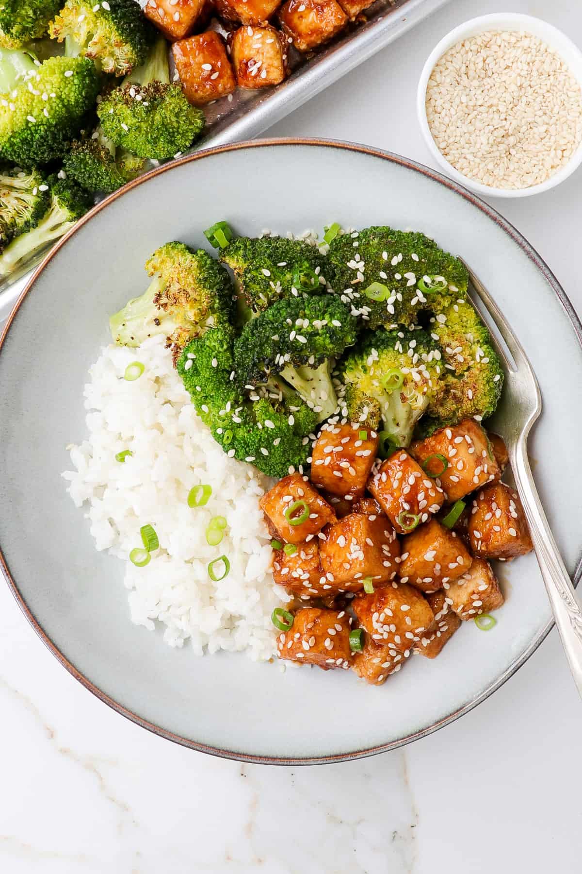 Honey garlic tofu in a bowl with roasted broccoli and rice. Sesame seeds and sheet pan on the side.