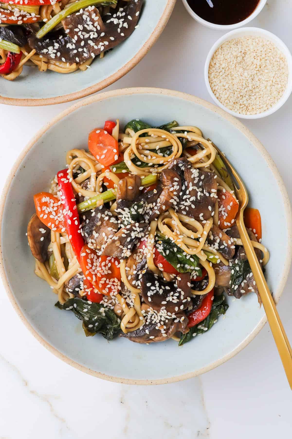 Teriyaki udon noodles with vegetables in a bowl with a gold fork. Sesame seeds sprinkled on top.