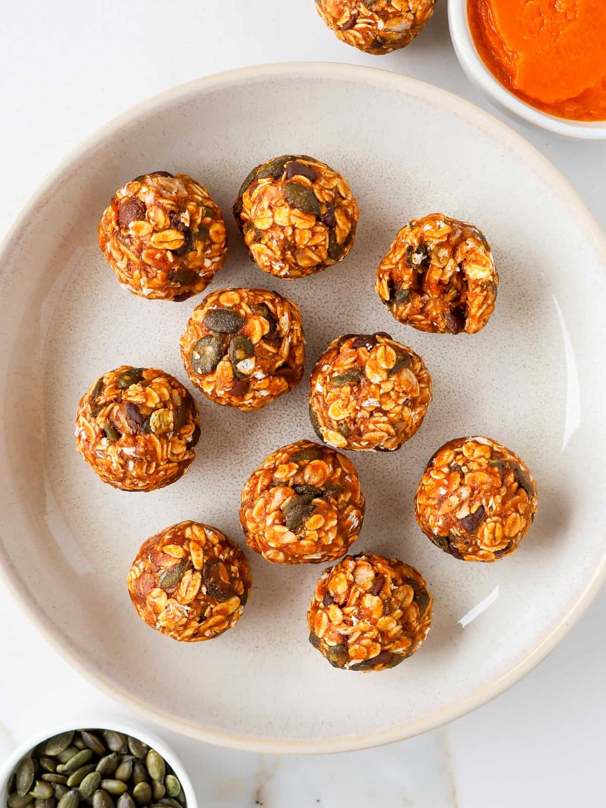 Pumpkin protein balls in a bowl with pumpkin puree and pumpkin seeds on the side. One ball has a bite.