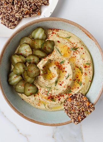 Hummus in a bowl topped with pickles, dill ad a seed cracker.