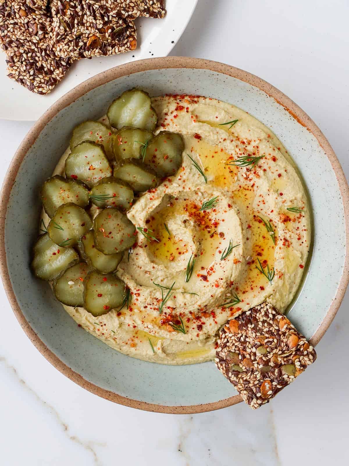 Hummus in a bowl topped with pickles, dill ad a seed cracker.