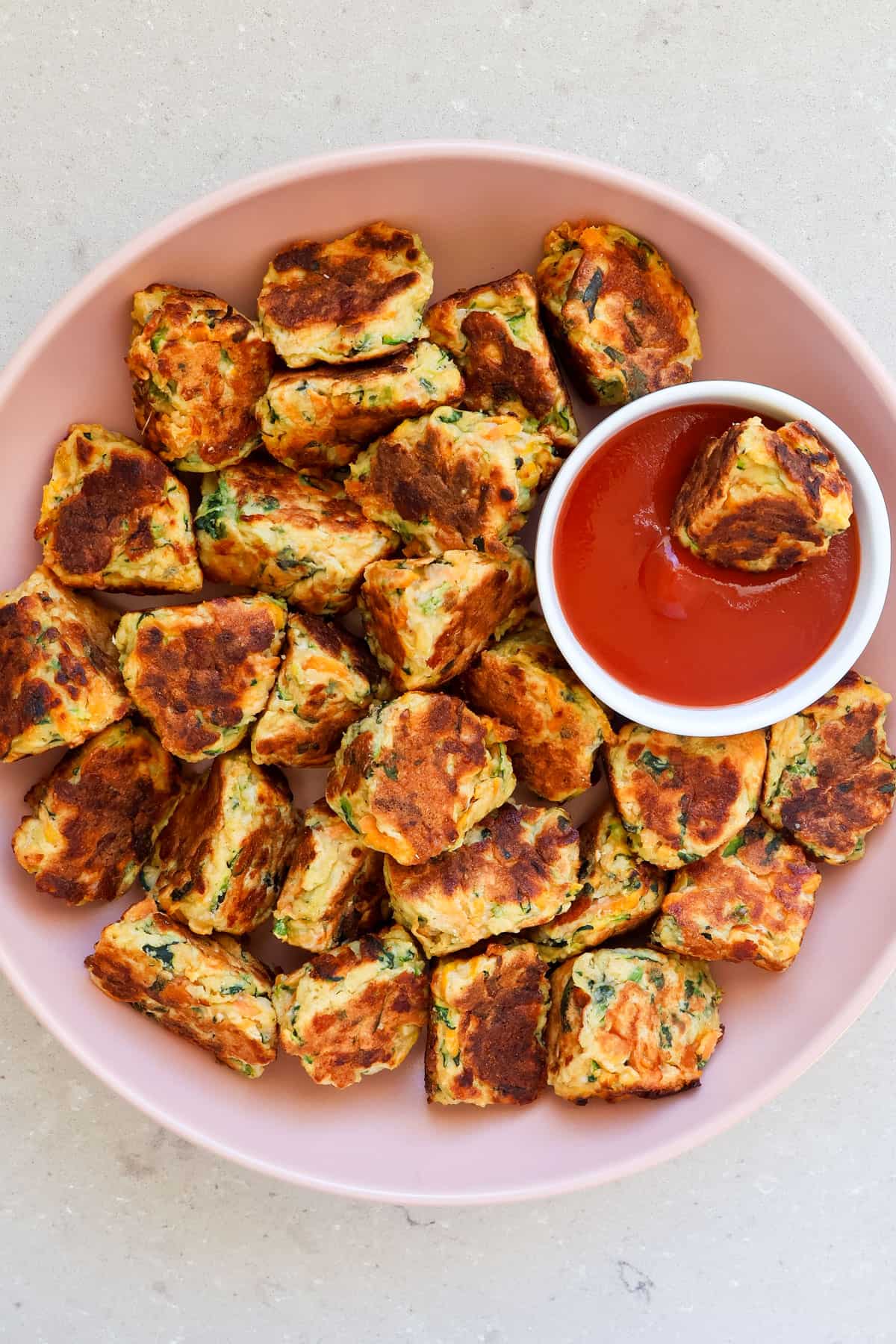 Ricotta veggie fritters in a bowl with one dipped in ketchup.