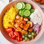 Chicken shawarma in a bowl with turmeric rice, cucumbers, tomatoes, pickles, onions and yogurt sauce.