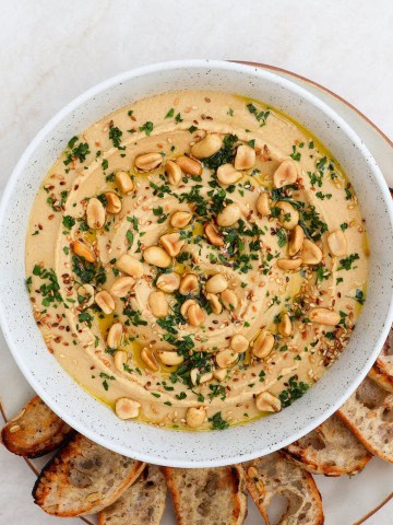 Peanut butter hummus in a bowl topped with peanuts, herbs, sesame seeds and crostini on the side.
