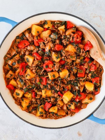 Ground beef, potatoes and peppers in a skillet with a wooden spoon.